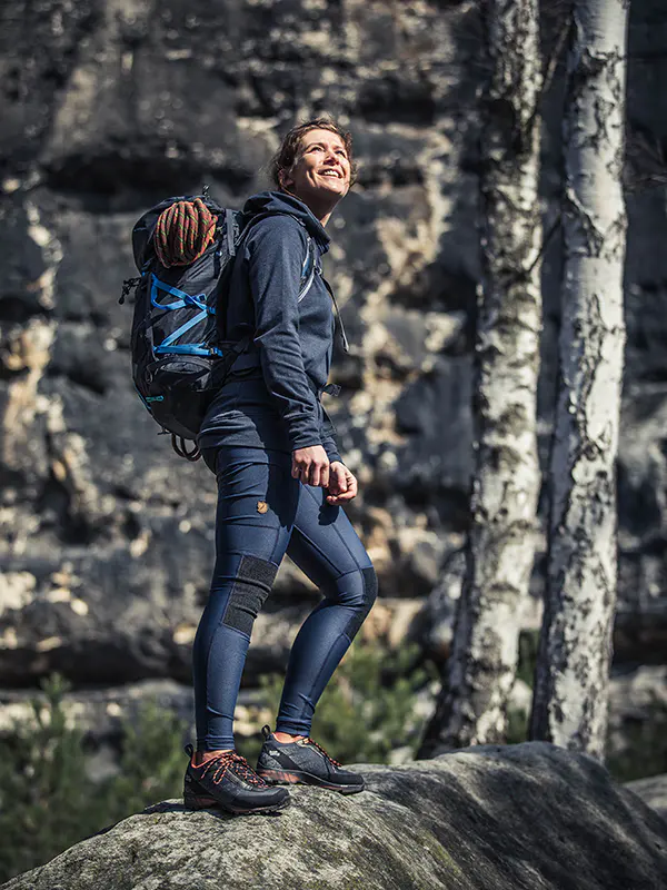 Een persoon in wandelkleding met een rugzak staat op een rots, glimlachend en omhoog kijkend, met op de achtergrond een rotsachtige klif en berkenbomen.